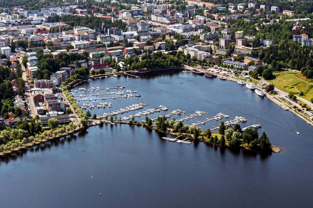 Aurinkoisella säällä otettu ilmakuva Lappeenrannasta. Kuvassa näkyy maisema Kaupunginlahden ja sataman yli kohti keskustaa. Etualalla sinistä Saimaata, taaempana keskustan rakennuksia ja vihreitä lehtipuita.