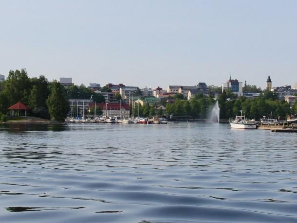 Lappeenrannan Kaupunginlahti Saimaalta keskustaan päin kuvattuna. Etualalla näkyy Saimaa ja pieniä laineita. Taaempana puita, veneitä sekä keskustan rakennuksia.