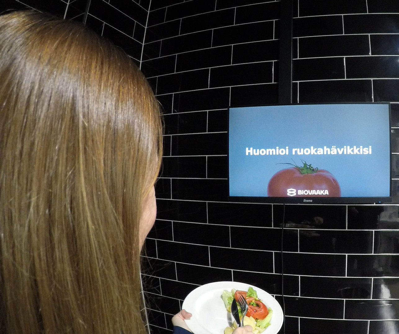 A person with a plate of food in front of a screen with the Biovaaka logo and the text Mind your food waste.