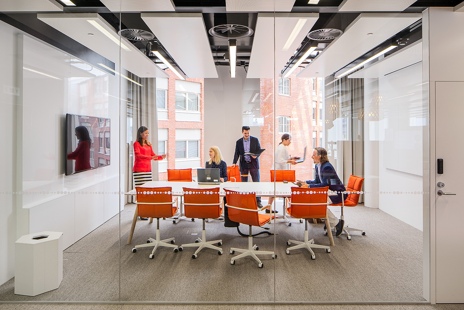 A conference room with a group of tables with orange chairs and tall windows in the background. People around the table, some sitting, some standing, some with laptops in their hands. Radiant heaters in the ceiling.